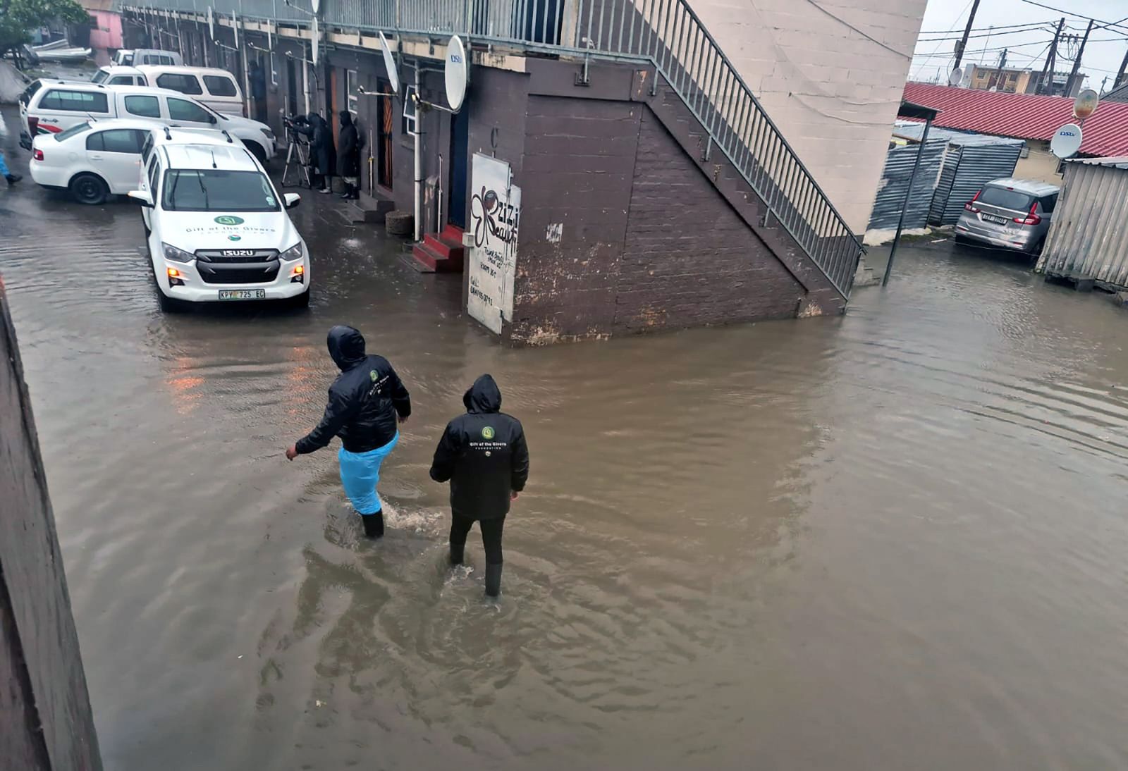 Flooded informal area with news camera crew walking through ankle-deep water in a parking lot.