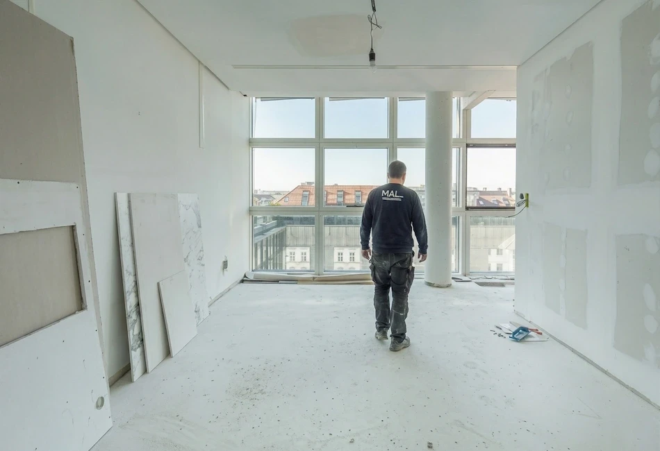 Handwerker betritt eine im Umbau befindliche Wohnung mit großen Fensterfronten, bereit für die Bodenverlegung