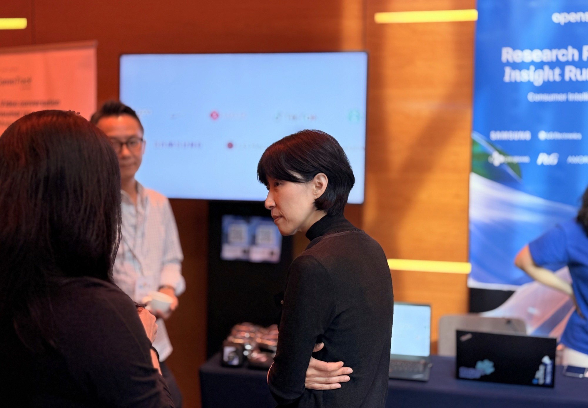 A group of people engaged in conversation at an event, with a presentation screen in the background.