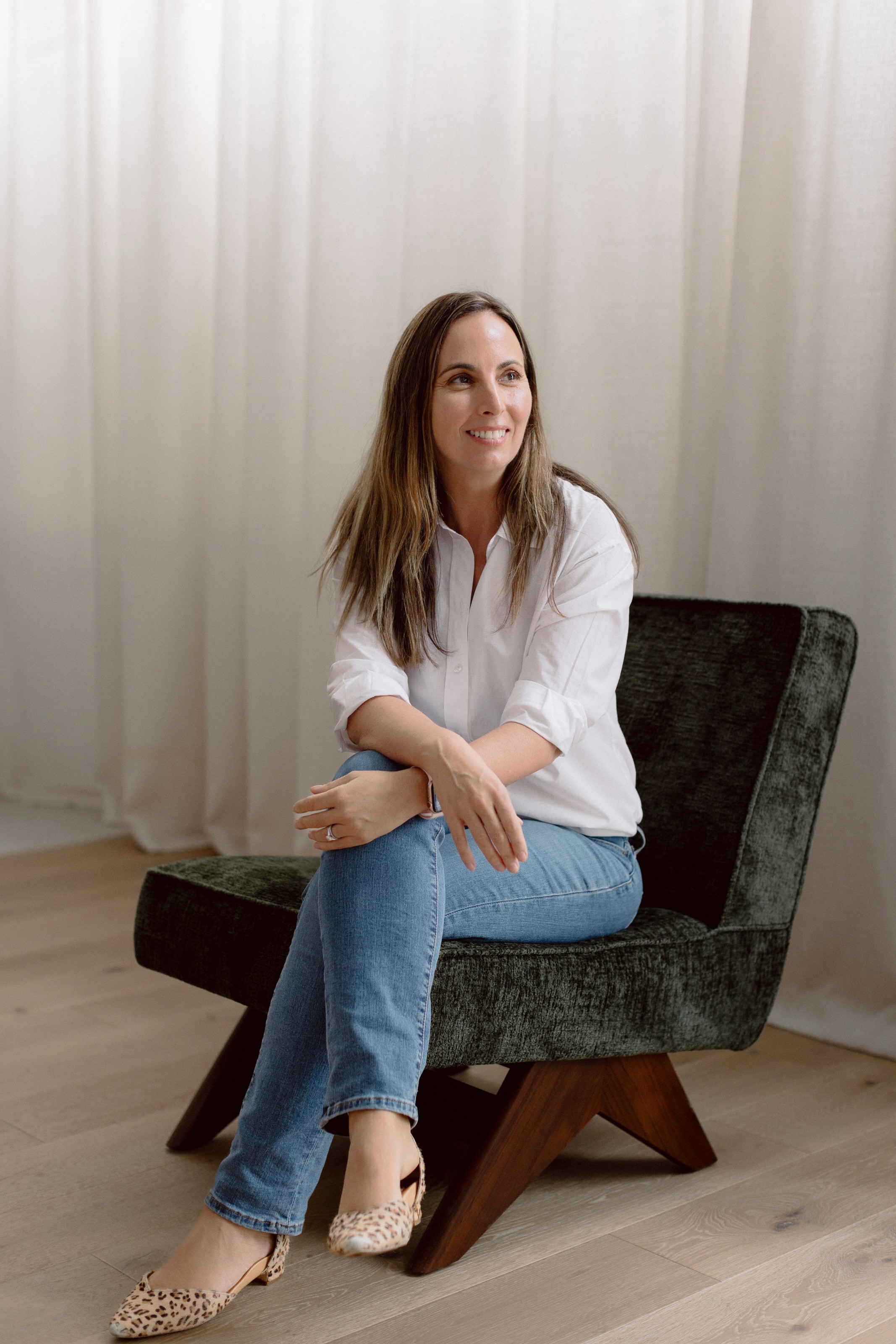 A woman smiling while sitting on a soft black chair, with a curtain softly draping in the background.