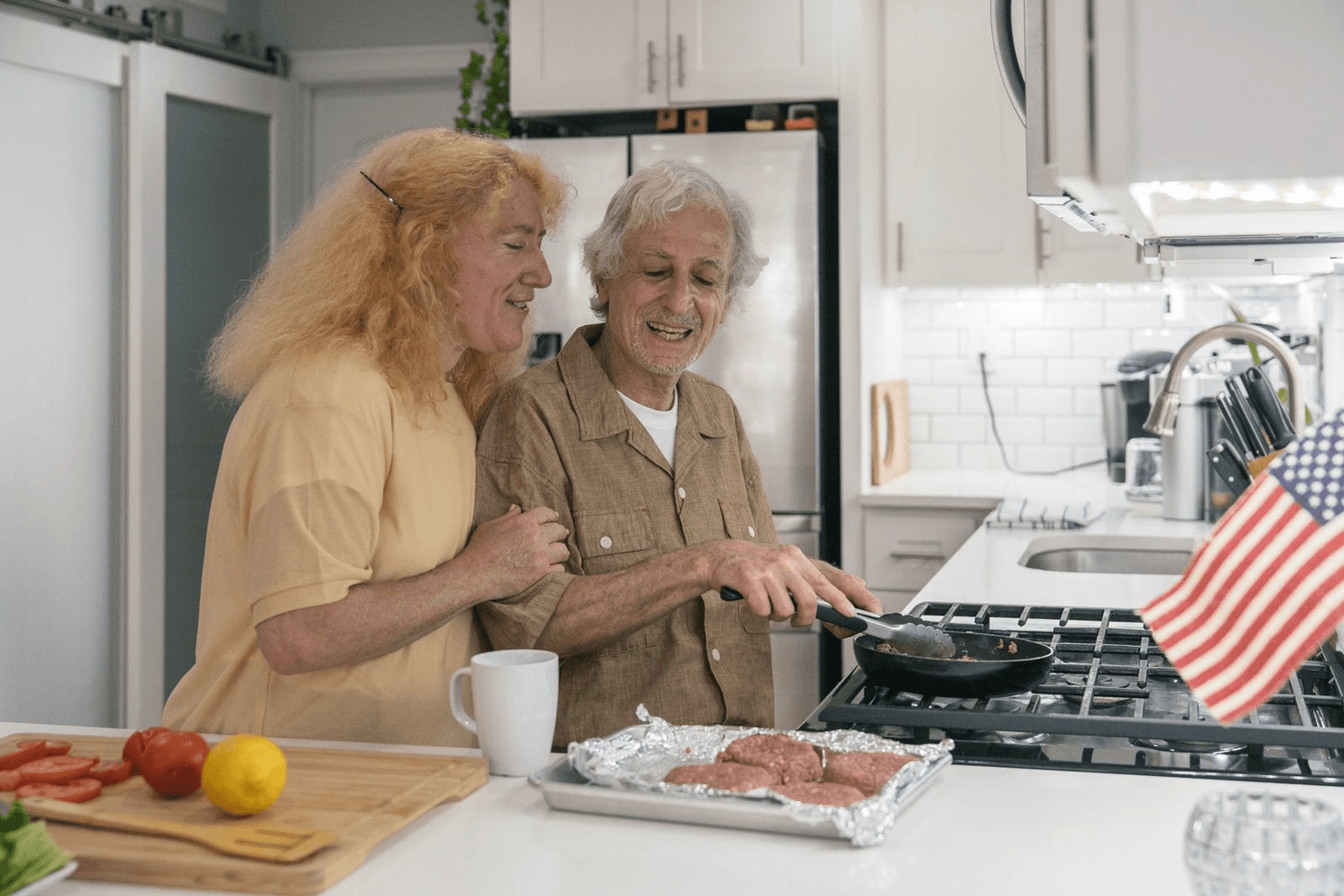 An older man and a helper cook together at the stove in a bright kitchen, both laughing.