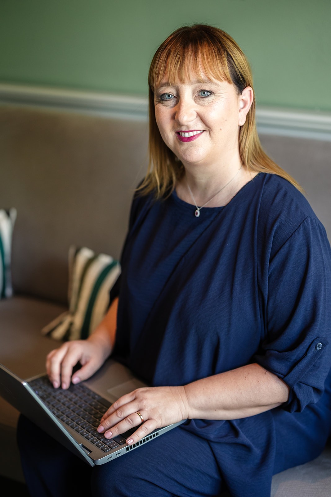 A young person with curly hair smiles at the camera while sitting in front of a laptop against a dark background.