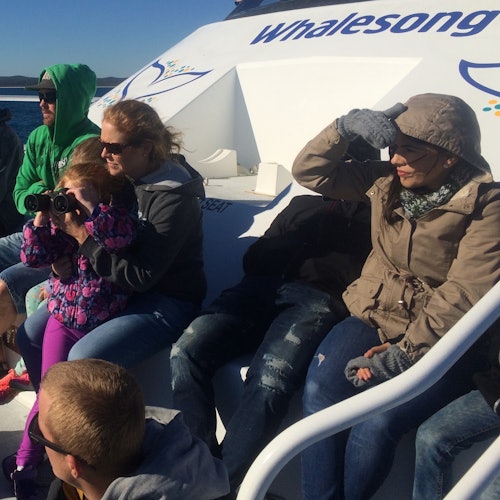 People on a boat are dressed warmly; one is using binoculars. The boat has "Whalesong" written on it.