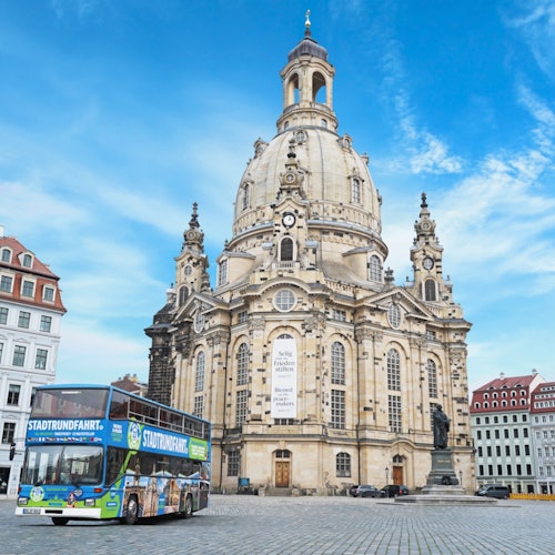 Barokkityylisen Dresdenin Frauenkirchen vieressä oleva kiertoajelubussi sinisen taivaan alla, taustalla historiallisia rakennuksia.