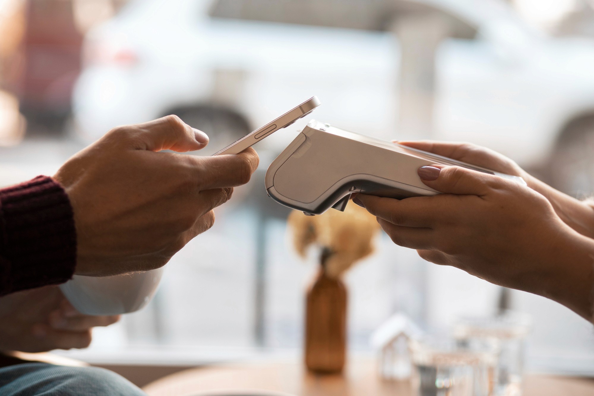 imagem que mostra de um lado a mão de um homem com o celular se aproximando da maquininha de cartão, que está sendo segurada pela mão de uma mulher, indicando que está sendo realizada uma venda e que o pagamento é por meio de aproximação do celular com o cartão online até a maquininha de cartão