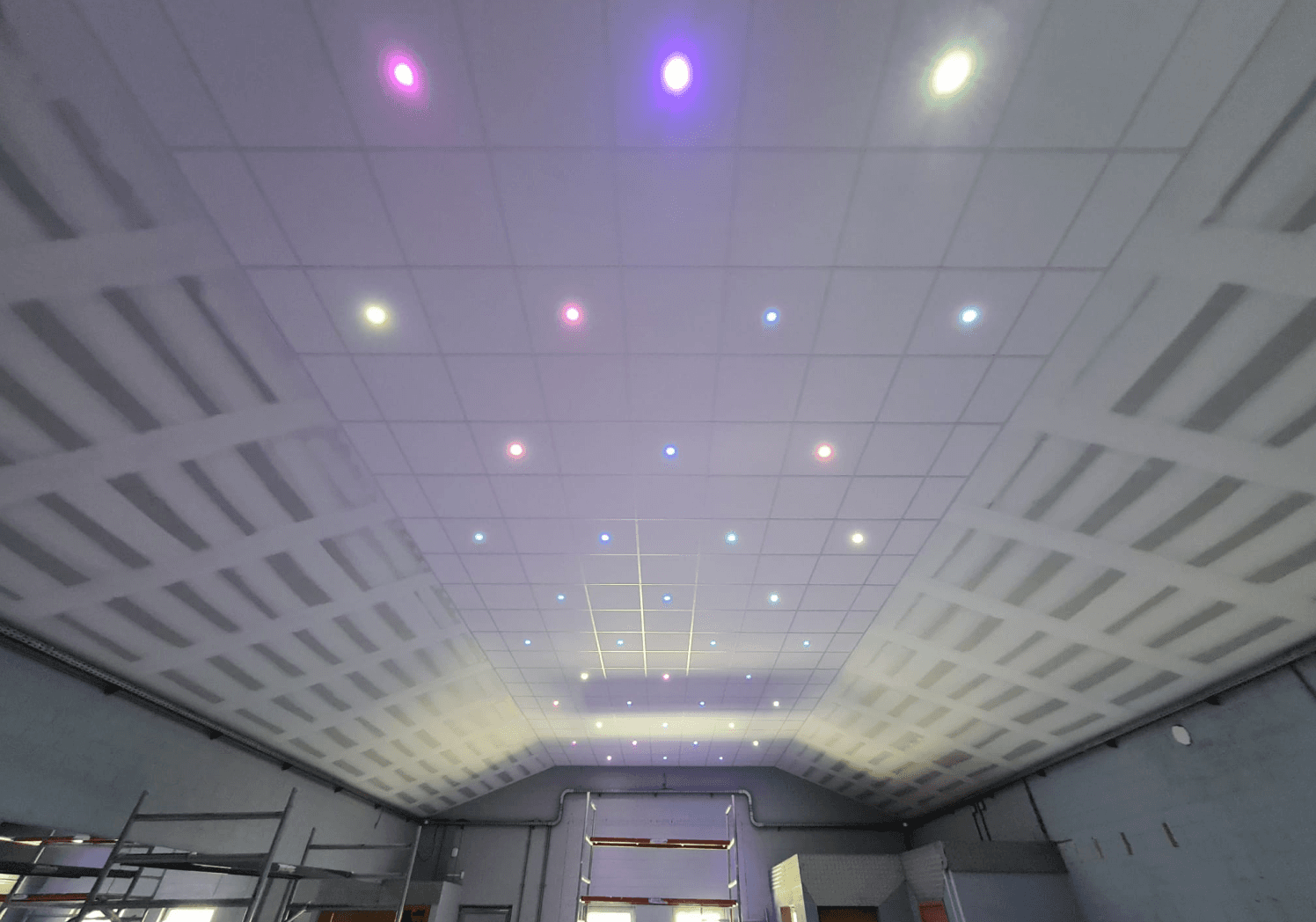 Plafond met talrijke kleine inbouwspots die verschillende kleuren uitstralen (roze, paars, groen, blauw), met een plafond van onafgewerkte gipsplaten of akoestische tegels, wat een gevoel geeft van renovatie of blootliggende structuur.