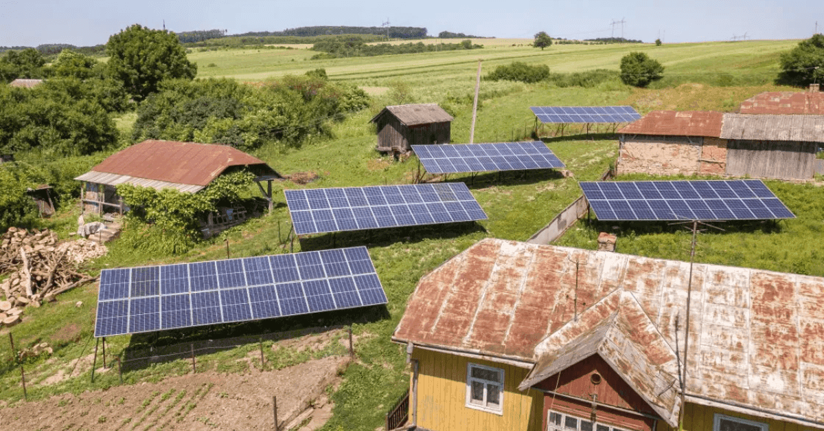 A rural community landscape featuring multiple ground-mounted solar panel arrays positioned around old farm buildings with rusted roofs, surrounded by green fields, trees, and open countryside under a bright sky.