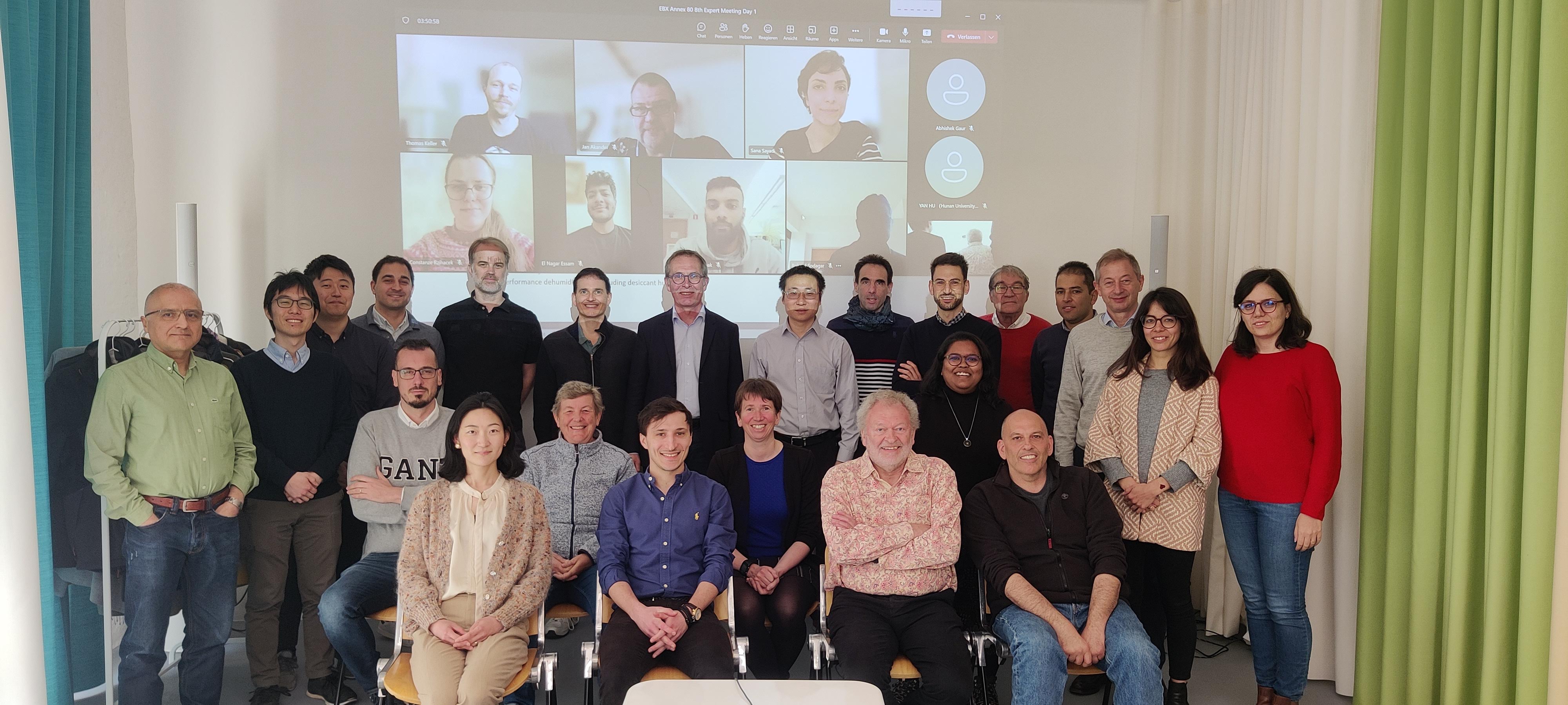 A group of around twenty people posing for a photo in a meeting room. Behind them, a screen shows additional participants joining the session online. The group includes individuals of diverse ages and backgrounds, standing and sitting together in front of light-colored curtains.