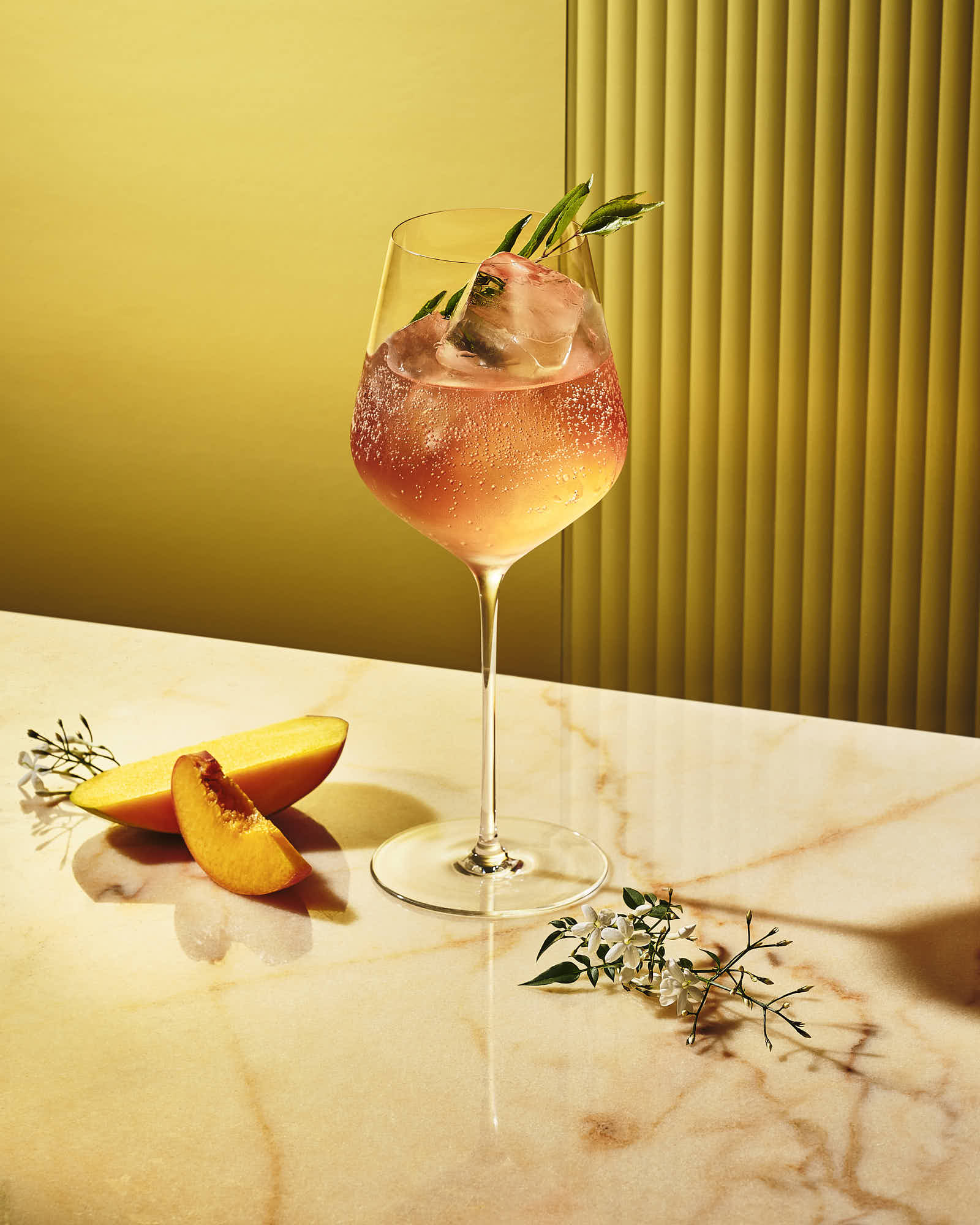 A cocktail glass with a pink drink garnished with a slice of fruit, resting on a light-colored surface with leaves.