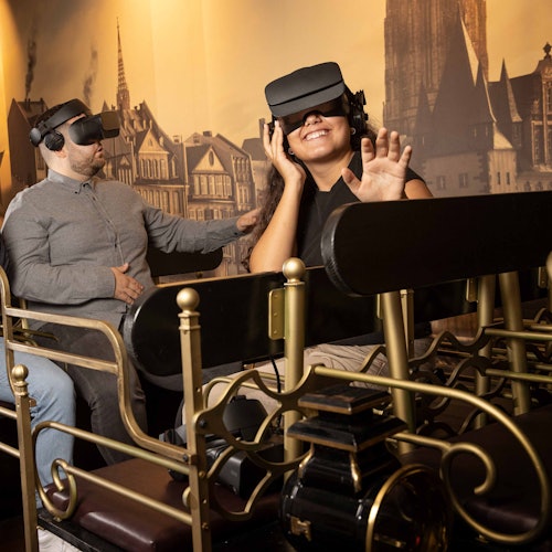 Two people wearing VR headsets sit in a ride with ornate rails, smiling and reaching out as if interacting with a virtual environment.