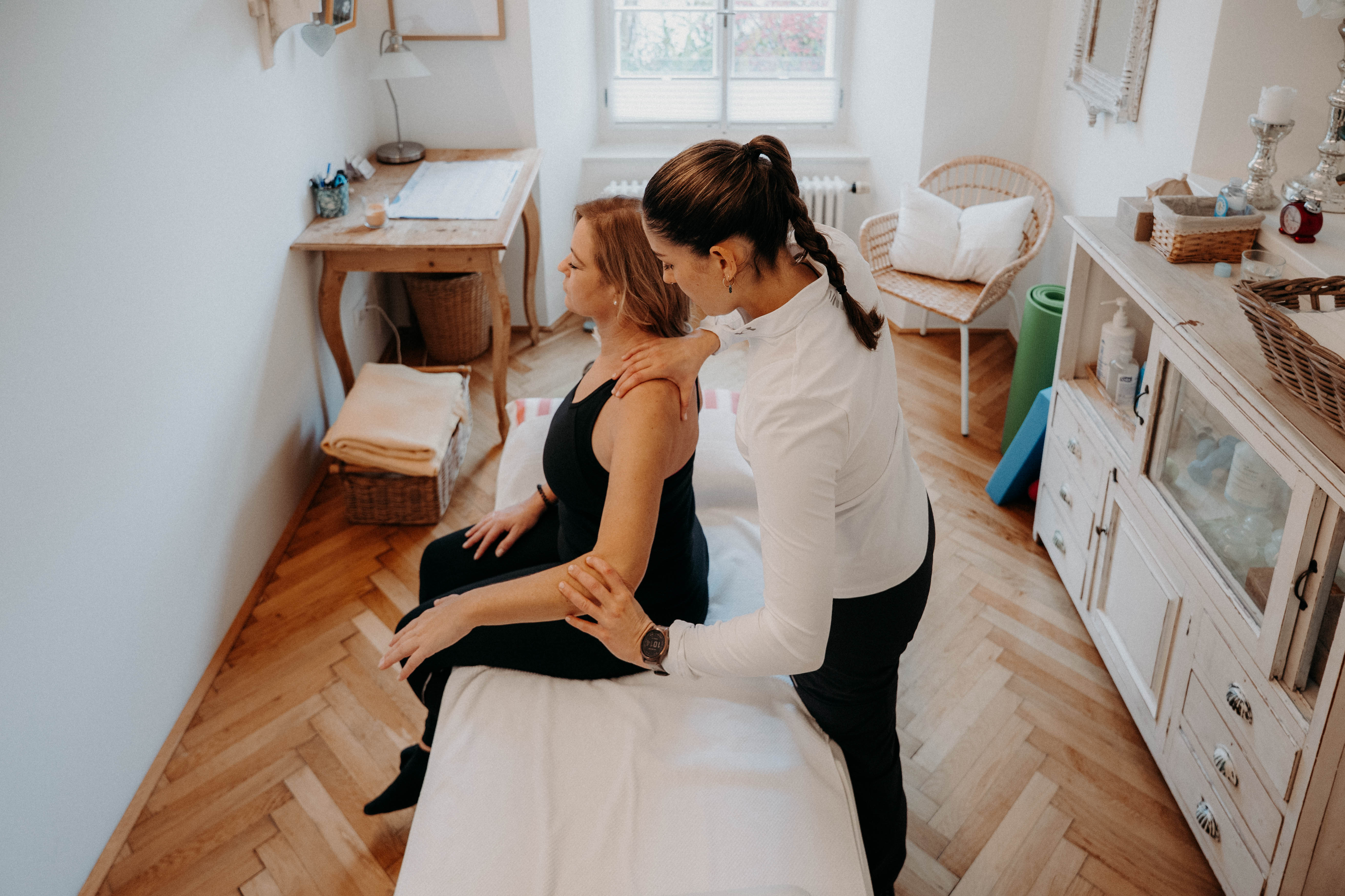 Physiotherapeutin behandelt Patientin, die auf der Behandlungsliege sitzt. Fokus auf Nackenmassage in hellem Raum mit Fenster.