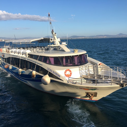 Bílý osobní trajekt jménem "Lord Byron" pluje klidnými modrými vodami pod čistou oblohou. Na pozadí jsou vidět hory.