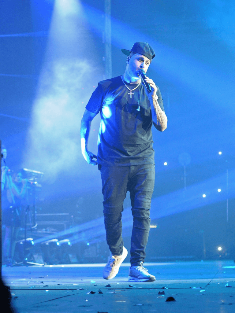Artista con gorra y crucifijo canta sobre escenario iluminado en azul; viste remera oscura, jeans y zapatillas claras.