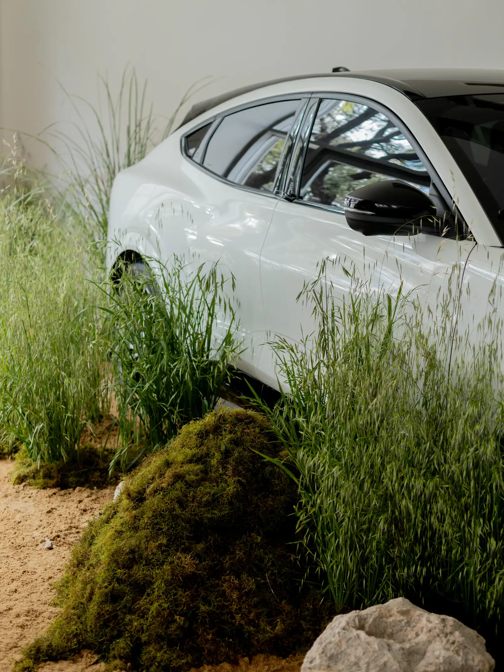 Vista lateral de auto blanco rodeado de hierbas y montículos de musgo en entorno minimalista interior.