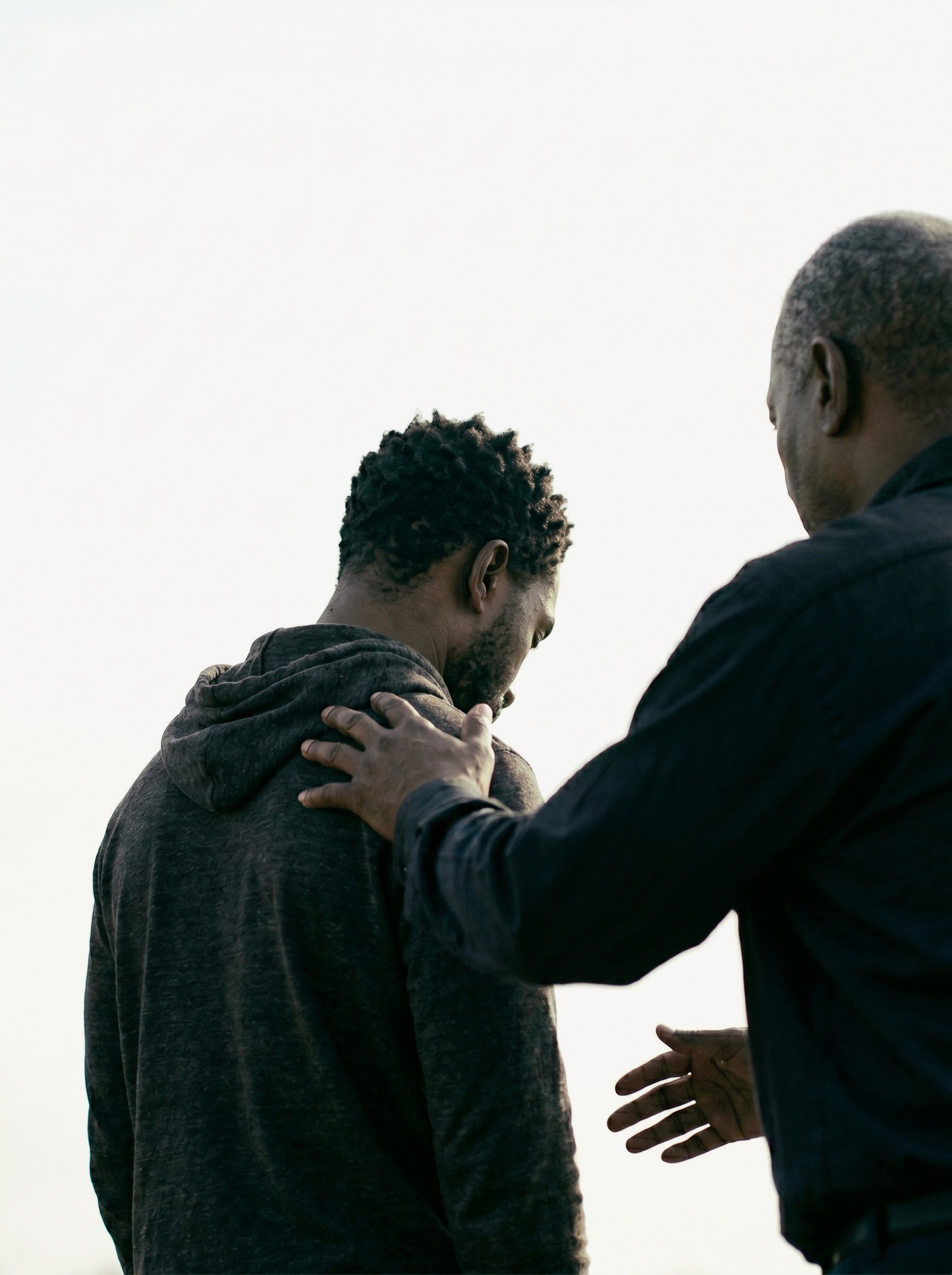 Two people are interacting outdoors; one person is gently placing a hand on the other's shoulder, conveying support and empathy on a clear day.