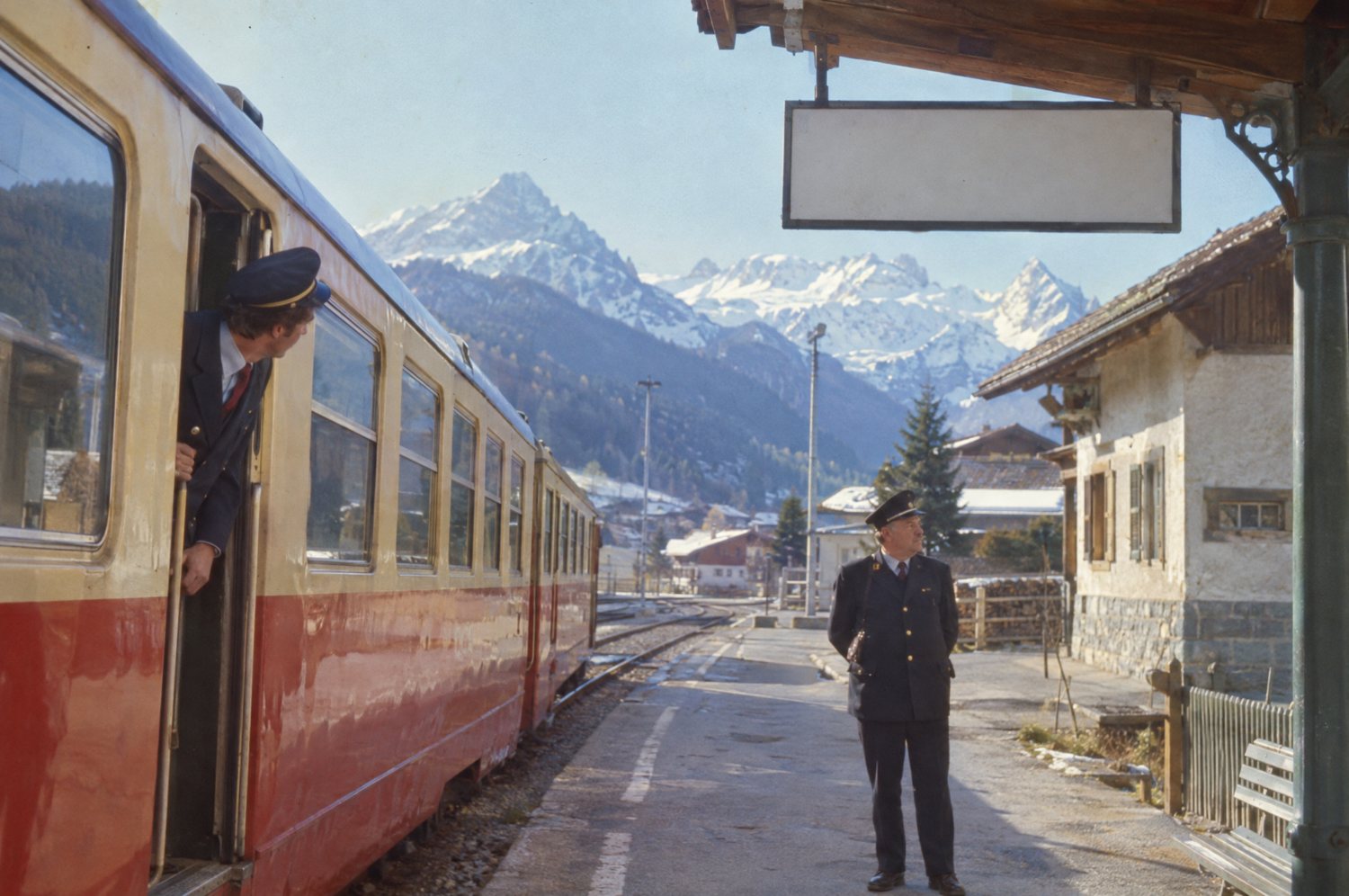 A 1970s colour slide of a small Alpine railway station: a stopped red-and-cream train, a conductor leaning out, a stationmaster on the platform, the station sign blank