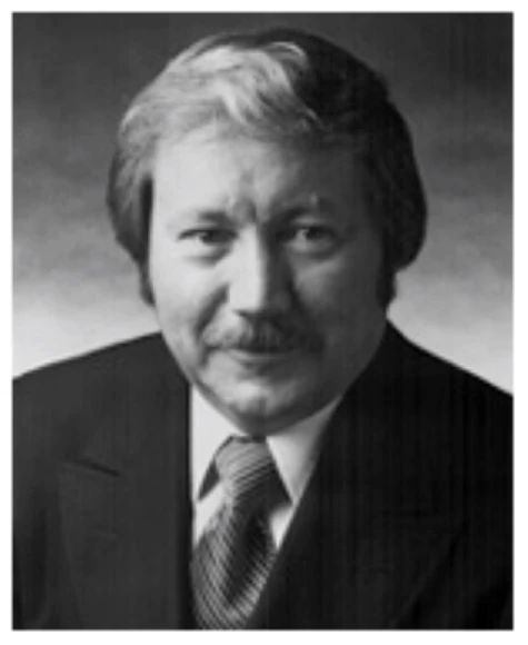 The image is a professional black and white portrait of a man in a suit and tie, sitting against a gradient background, conveying a formal and classic style.