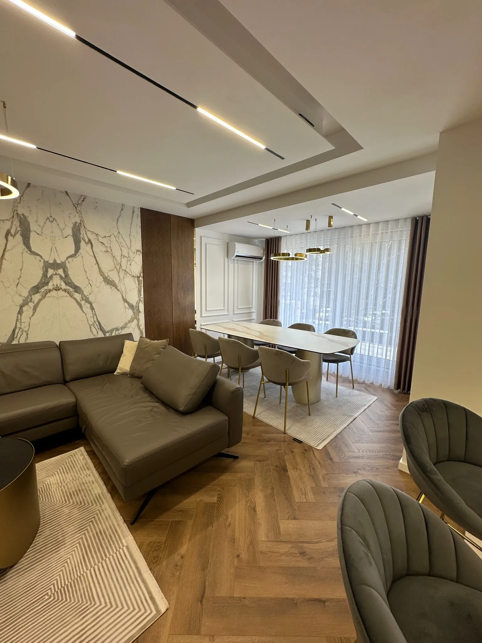 Modern living and dining area in a Gostivar apartment featuring custom interior design by Terraplan Partners, with marble accent walls, warm wood flooring, contemporary lighting, and a tailored furniture layout.