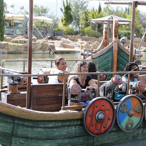 Ihmiset nauttivat ajelusta merirosvoteemaisella veneajelulla huvipuistossa, taustalla järvi ja vehreä luonto.