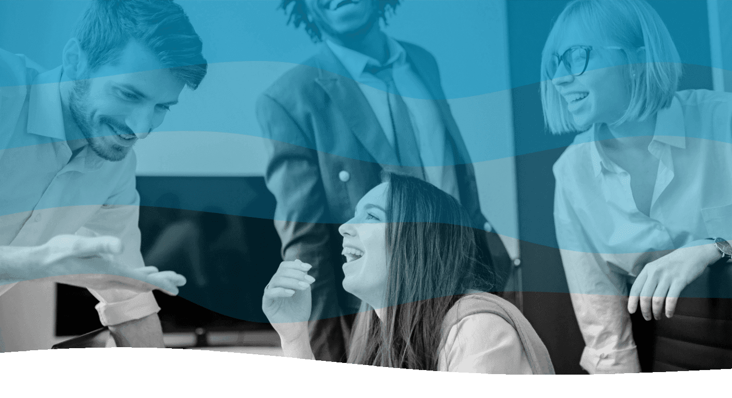 A group of colleagues in an office environment are enthusiastically discussing around a table, with one person seated and others standing, all sharing smiles and laughter, indicating a positive and collaborative work atmosphere.