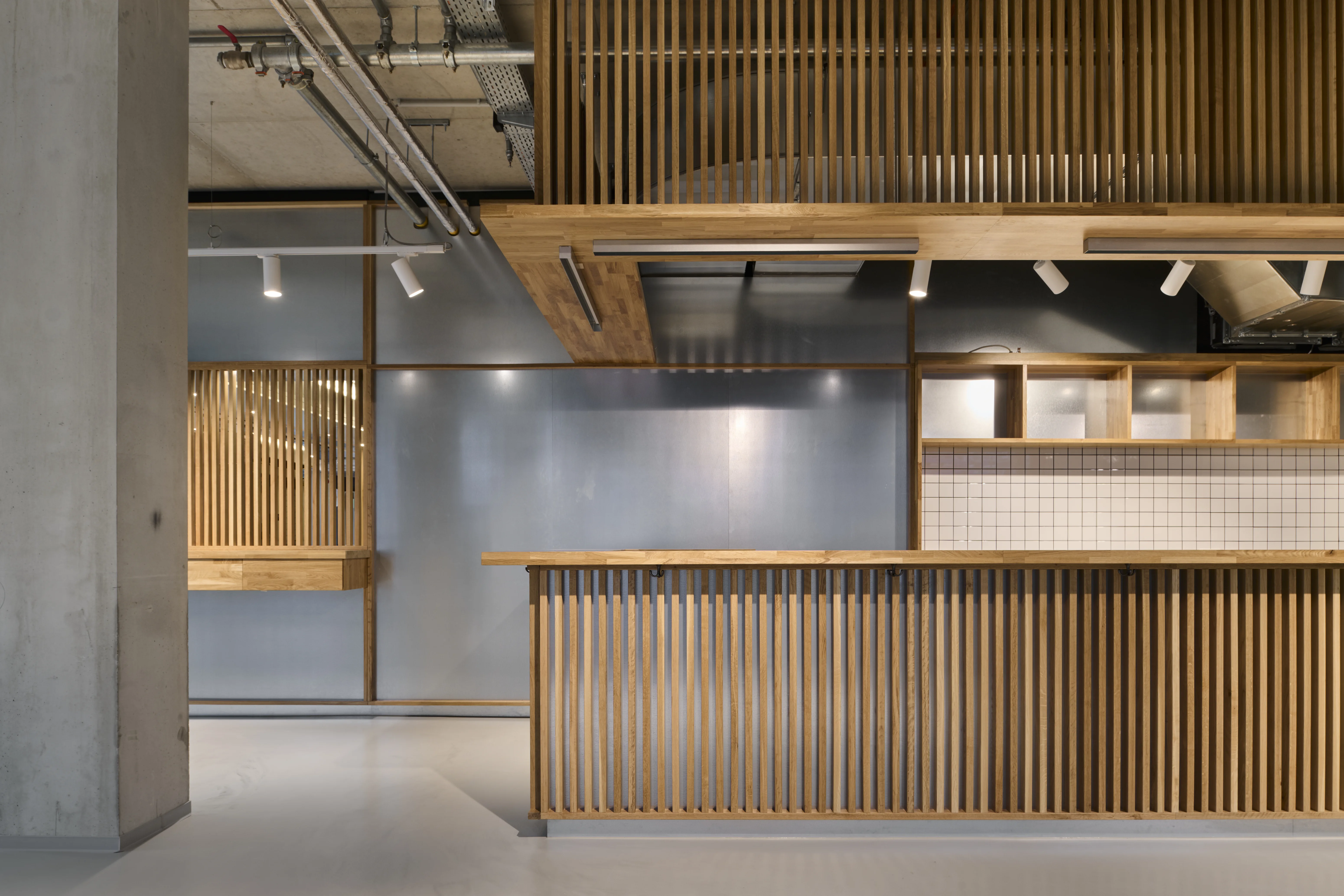 Wooden slat-fronted counter at PARKER BOWLES & FRIENDS with oak top, white tiled niche with open shelving and brushed metal wall panels.