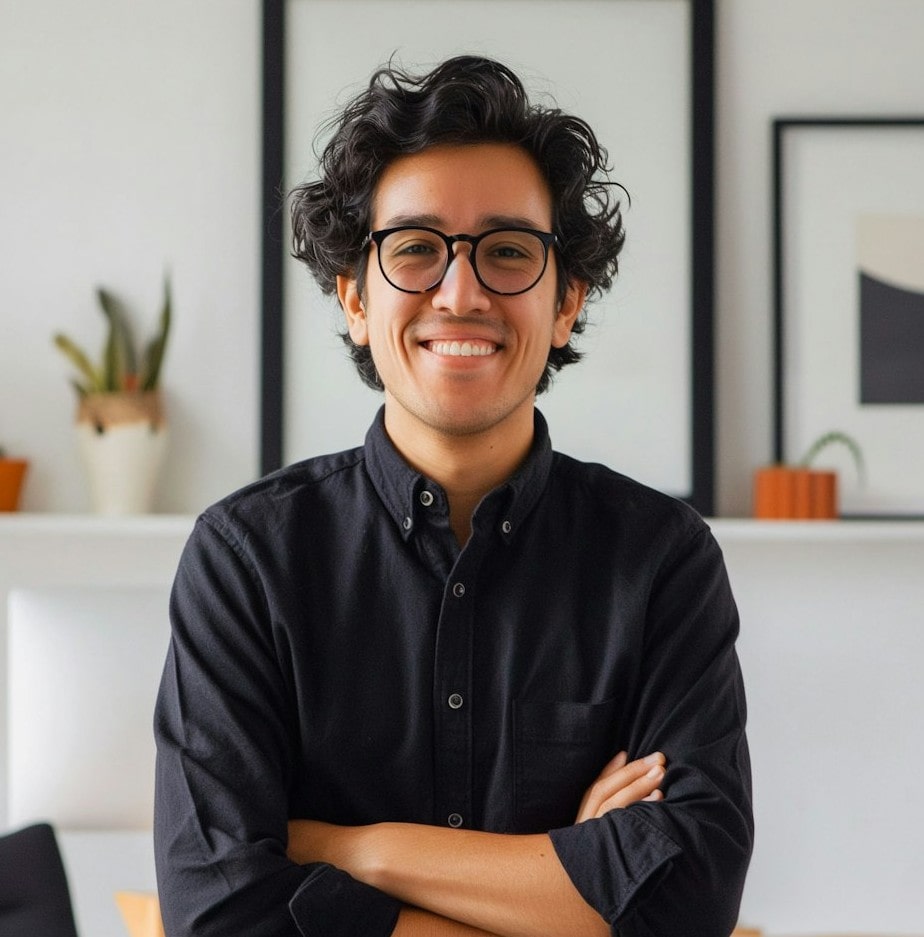 A man wearing glasses and a black shirt stands against a plain white wall.