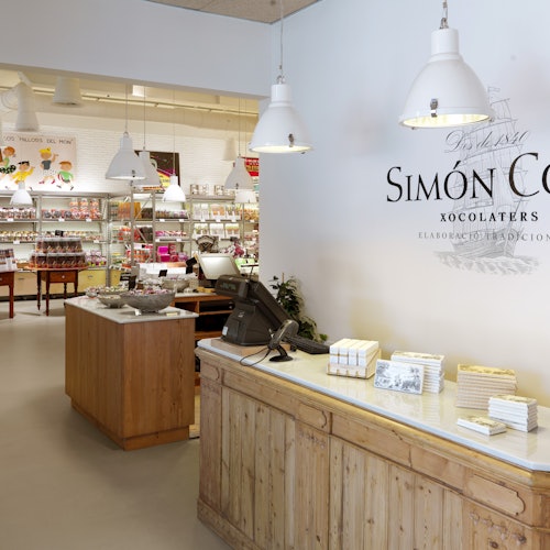 Un interior de botiga amb un taulell de fusta, una caixa registradora, llums penjants, prestatges amb productes de xocolata i un rètol que diu "Simón Con."