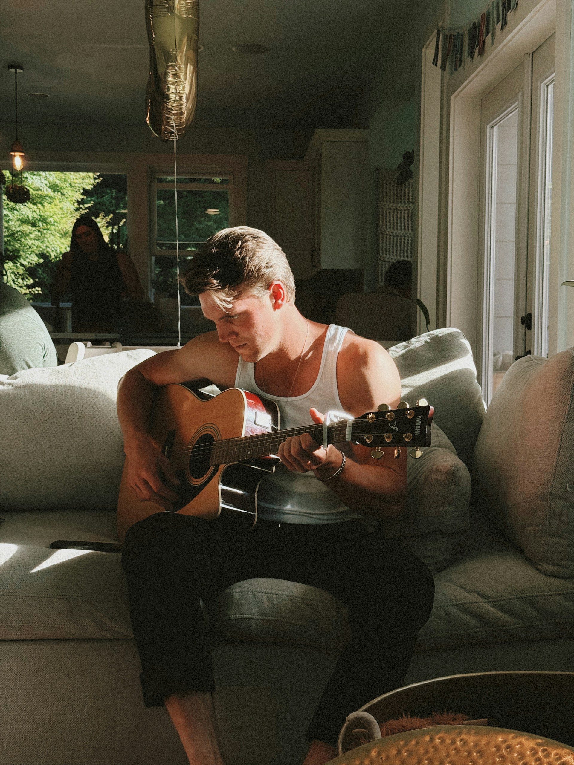 A candid photograph of a young man in a white tank top and black pants sitting on a couch, focused on playing an acoustic guitar in a sunlit room.