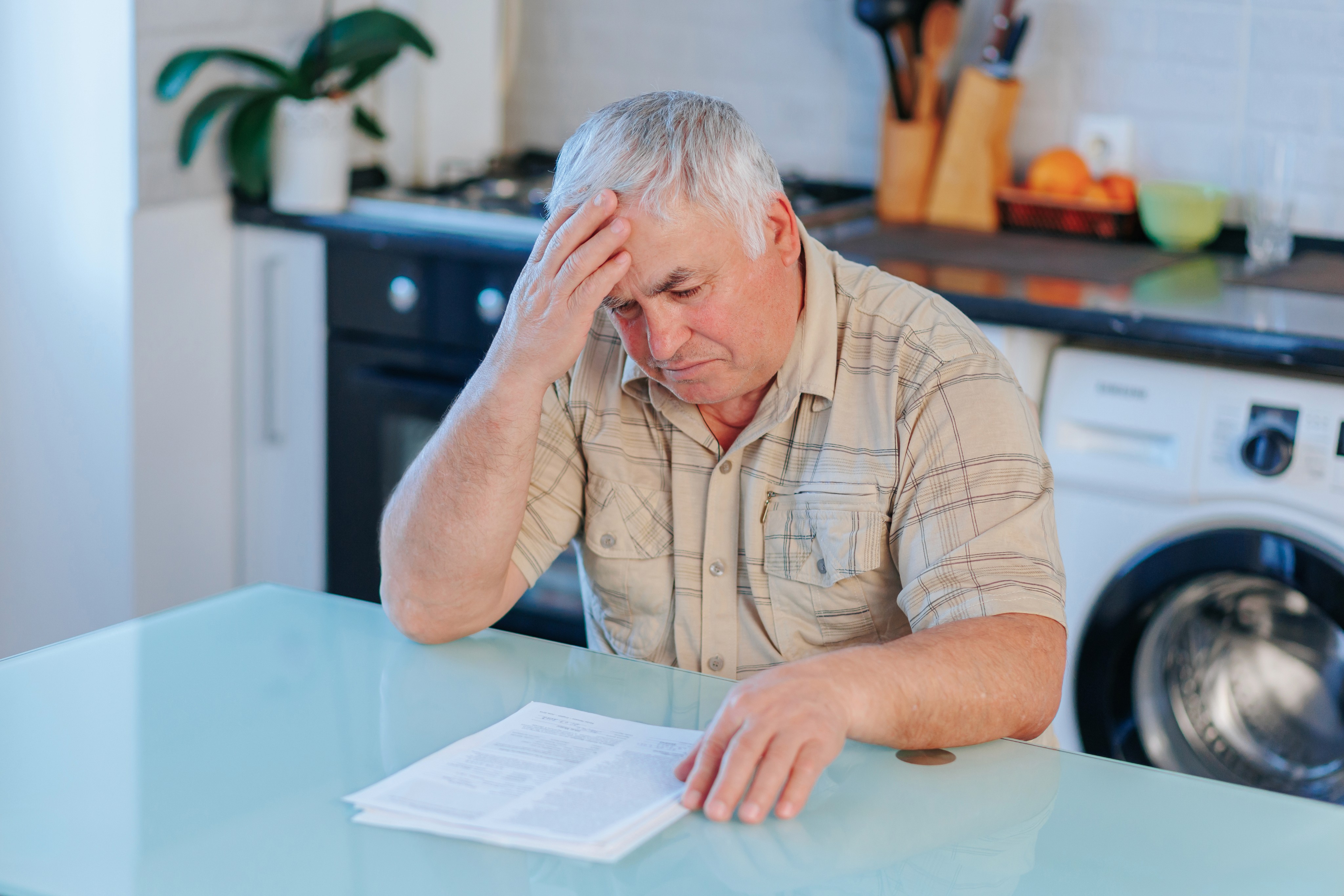 Elderly man in a kitchen, looking worried while holding a paper, sitting beside a laptop. His expression conveys concern and contemplation.