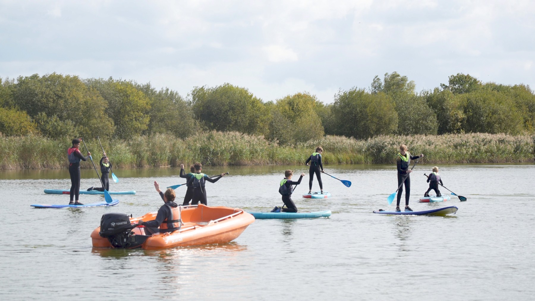 A group of people paddleboarding on a calm lake, accompanied by an orange motorboat. The scene is set against a backdrop of lush green trees.