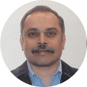 A person with short dark hair and a mustache is wearing a striped shirt and a dark jacket against a plain light background.