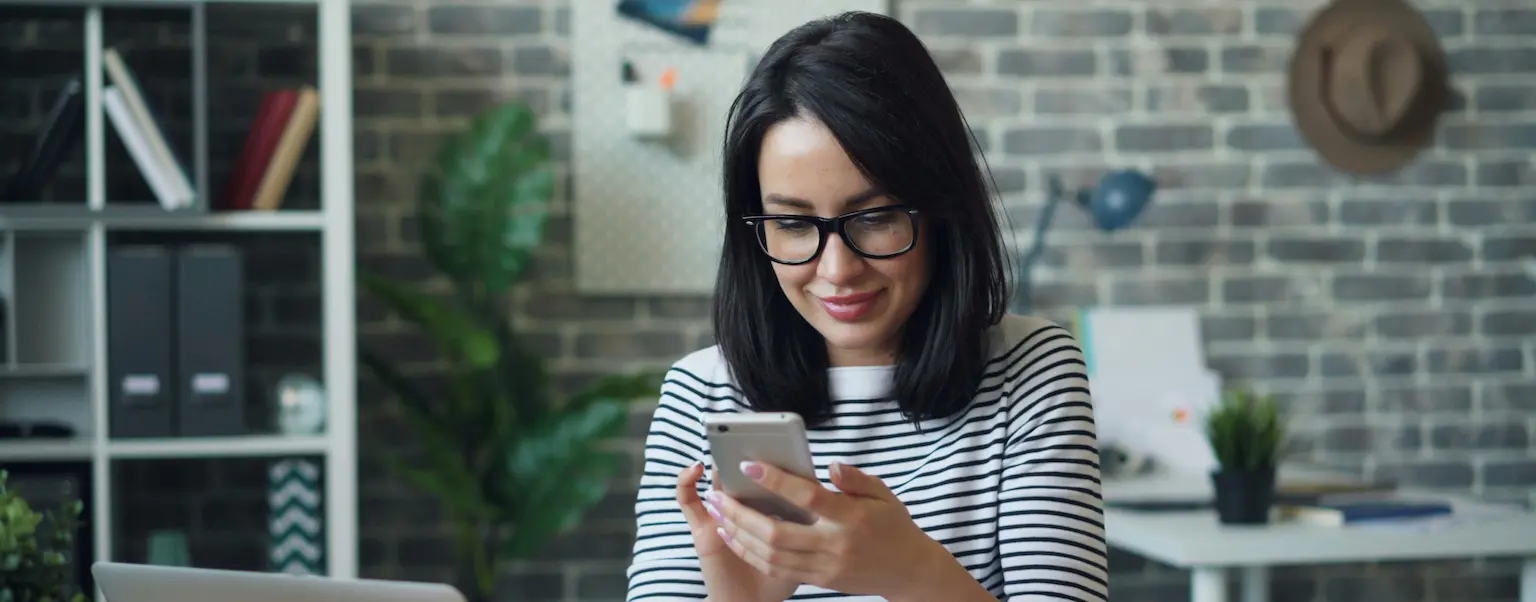 A woman sitting at a desk smiling while using her phone, with a laptop and home office setting in the background.