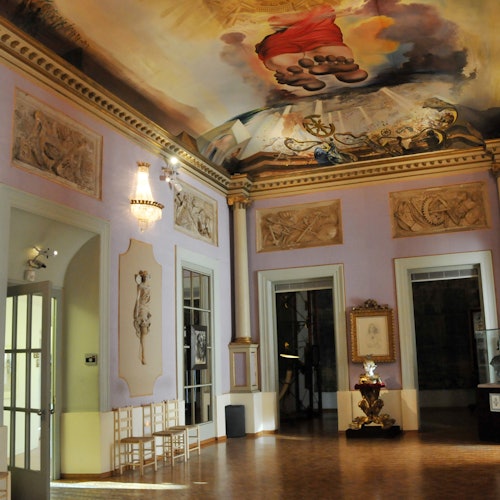 חדר מפואר עם ציורים קלאסיים, נברשות ואמנות תקרה דקורטיבית. כמה כיסאות ויצירות אמנות ממוסגרות נראים לעין.