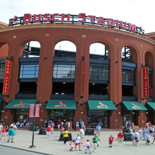 Crowd outside Busch Stadium entrance with green awnings and "Team Store" signs under the stadium's arched facade.