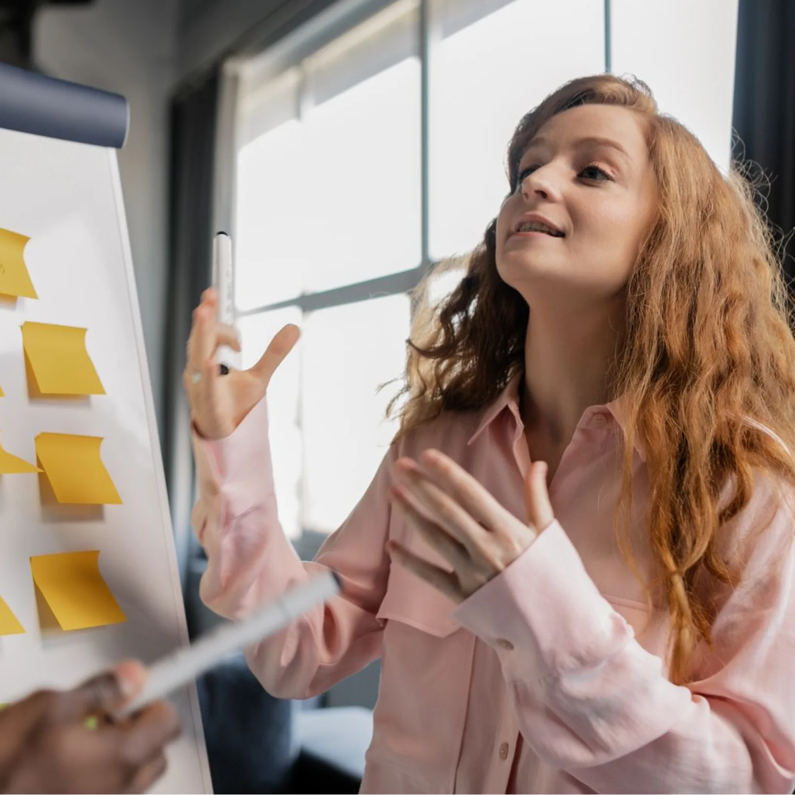 A woman with long, wavy reddish-brown hair giving an enthusiastic presentation next to a white flipchart. She is wearing a light pink button-down shirt and gesturing actively with both hands as she explains an idea. Several yellow sticky notes are attached to the flipchart. In the bottom left foreground, a Black colleague holds a marker, pointing toward the board during the collaborative discussion. The setting is a bright office with a large window in the background.
