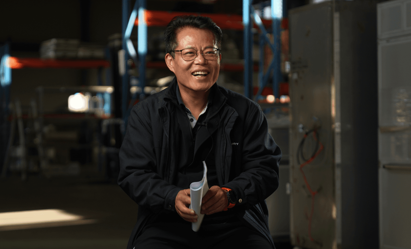 A modern bedroom features a cozy bed with white pillows, a striped rug, and a stone wall with a fireplace. Art with blue stones adds flair, while soft light creates a serene atmosphere.