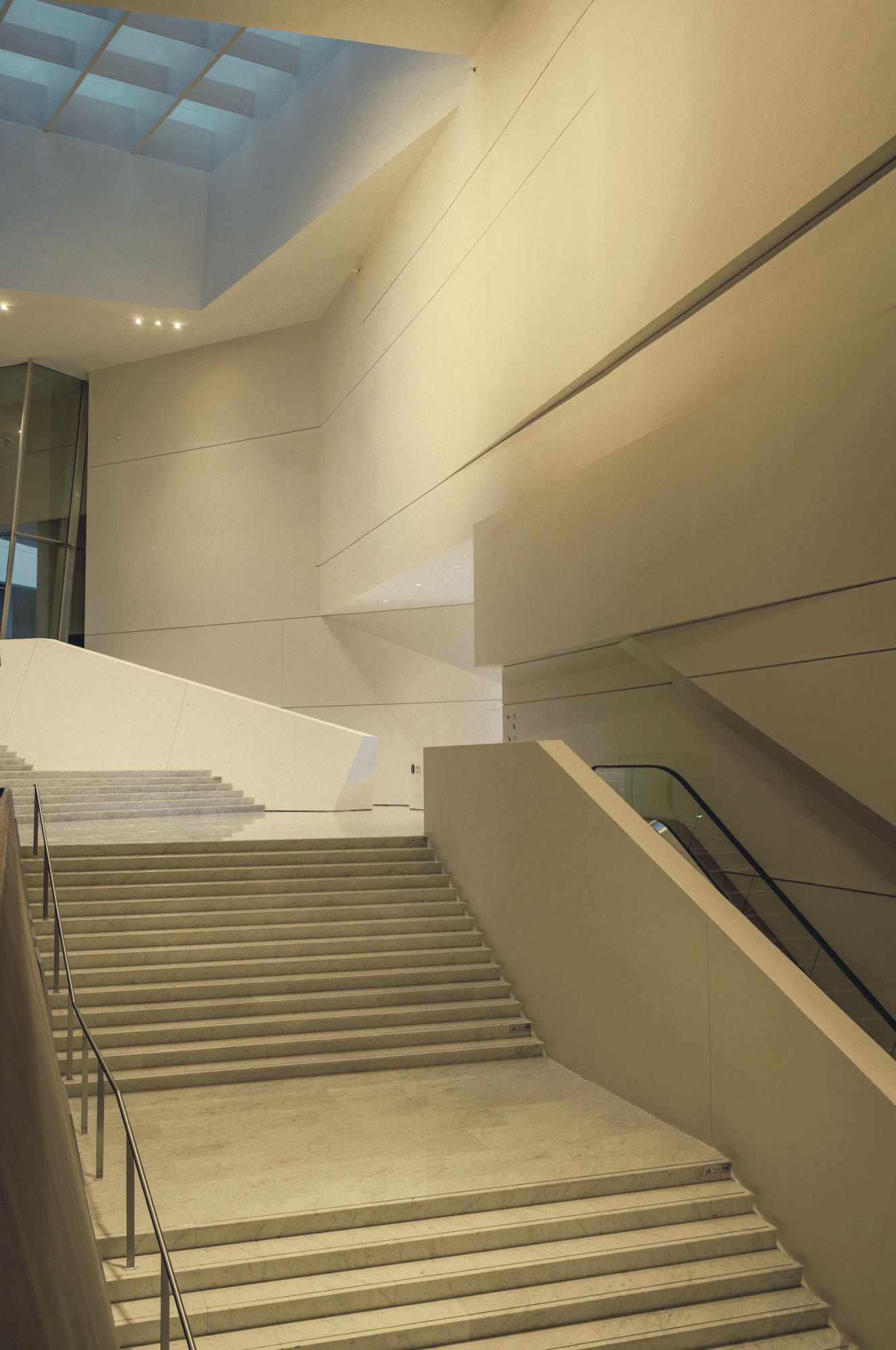 A wide stone staircase beside an escalator, rising toward the skylight