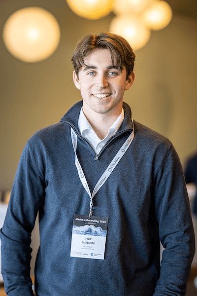 A young man in a dark sweater smiles at the camera, with soft lighting and blurred backgrounds. He wears a name tag.