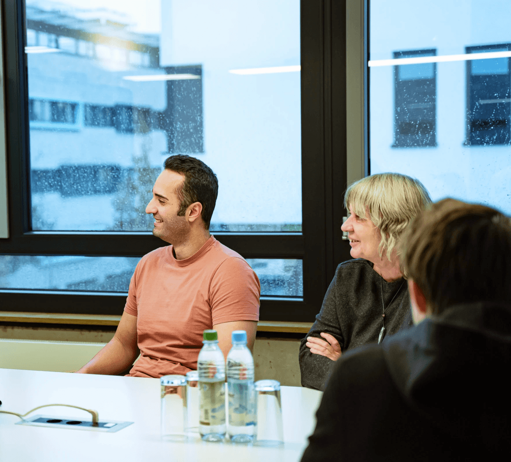 Mitarbeitende der kba Architekten und Ingenieure GmbH sitzen im Besprechungsraum vor großen Fenstern an einem Tisch, sprechen und lachen gemeinsam; im Vordergrund stehen Wasserflaschen und Gläser.