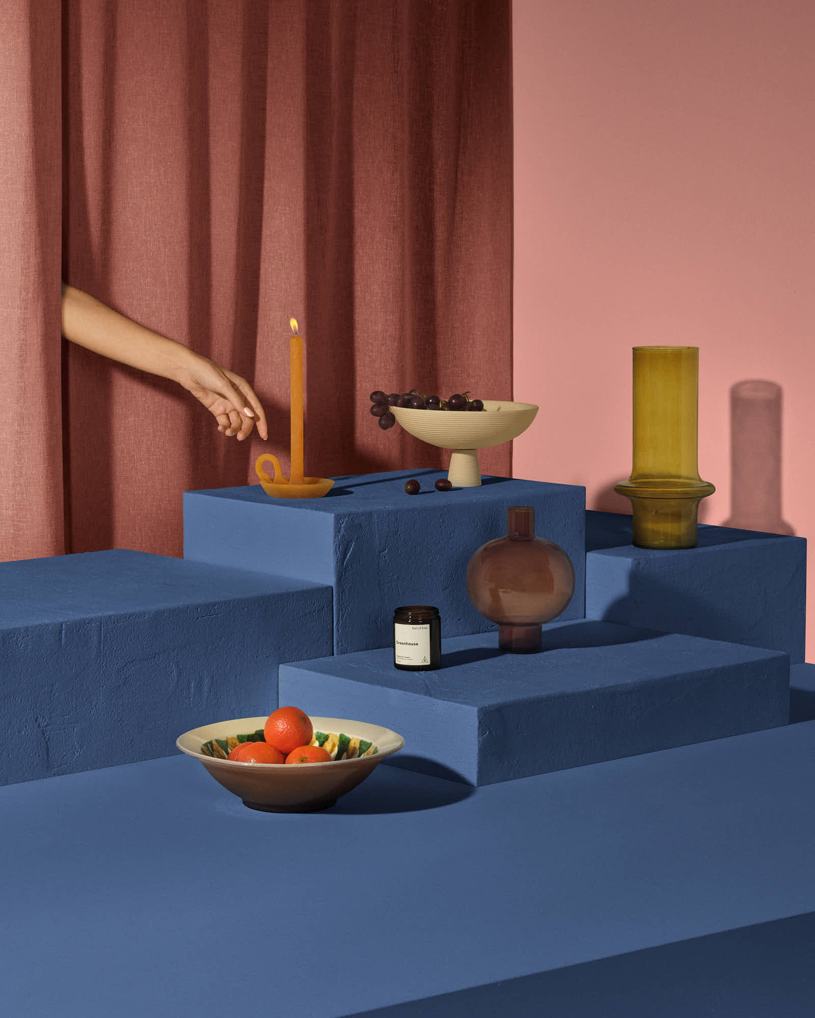 Colorful modern still life with candles, bowls, hand, and fruit on blue platforms against curtain backdrop.