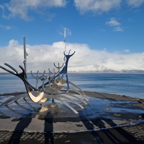 Escultura metálica que se asemeja a un barco con estructuras altas y curvas, situada contra un fondo de agua y montañas cubiertas de nieve bajo un cielo azul.