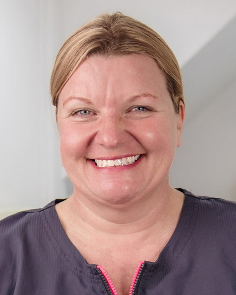 A portrait of Anne, a Dental Hygienist at Cricklade Dental Practice, smiling and wearing a dark grey scrub top with a pink zipper pull and yellow embroidered text that reads "ANNE" on the left and "Cricklade Dental Practice" on the right. She is standing in a dental surgery with a dental light visible in the background.
