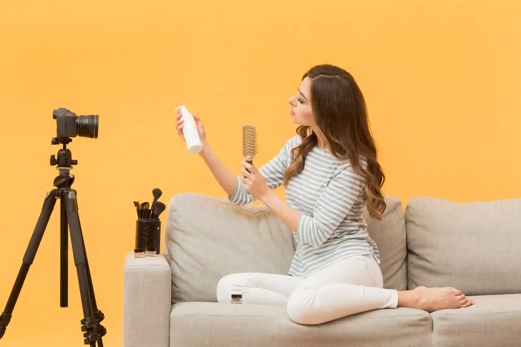 Young woman recording a video reviewing a cosmetic product and holding a hairbrush on a sofa.