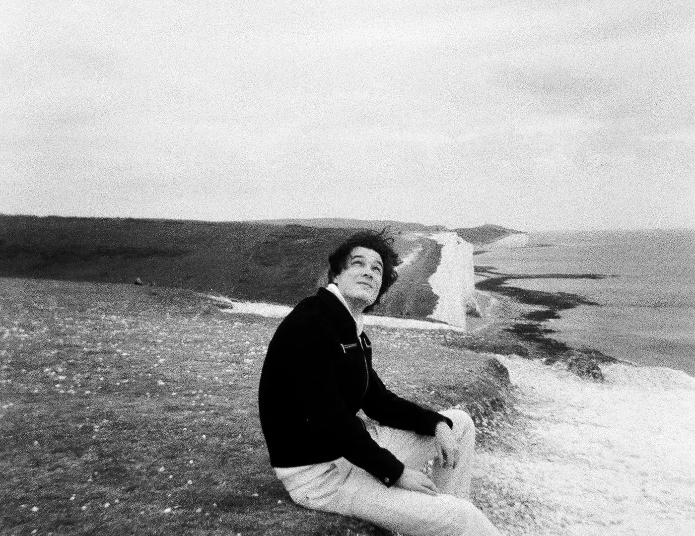 Black-and-white photo of Jonas Parna sitting on a grassy clifftop above white chalk cliffs and the sea