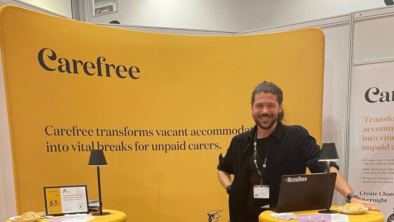 Smiling Joey Ceunen standing at a Carefree exhibition booth with a yellow backdrop that reads 'Carefree transforms vacant accommodation into vital breaks for unpaid carers.' The booth features a laptop, small lamps, printed materials, and Carefree branding.