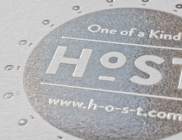 A close-up view of a circular metal plate with a textured surface. The plate features the text "One of a Kind" at the top, followed by the prominent letters "HOST" in a larger font. Below this, a thin horizontal line separates the upper text from the website address "www.h-o-s-t.com". The plate has a series of small circular indentations along its outer edge.