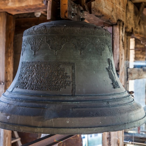 An aged, ornate bell with inscriptions hangs from wooden beams in a rustic setting.