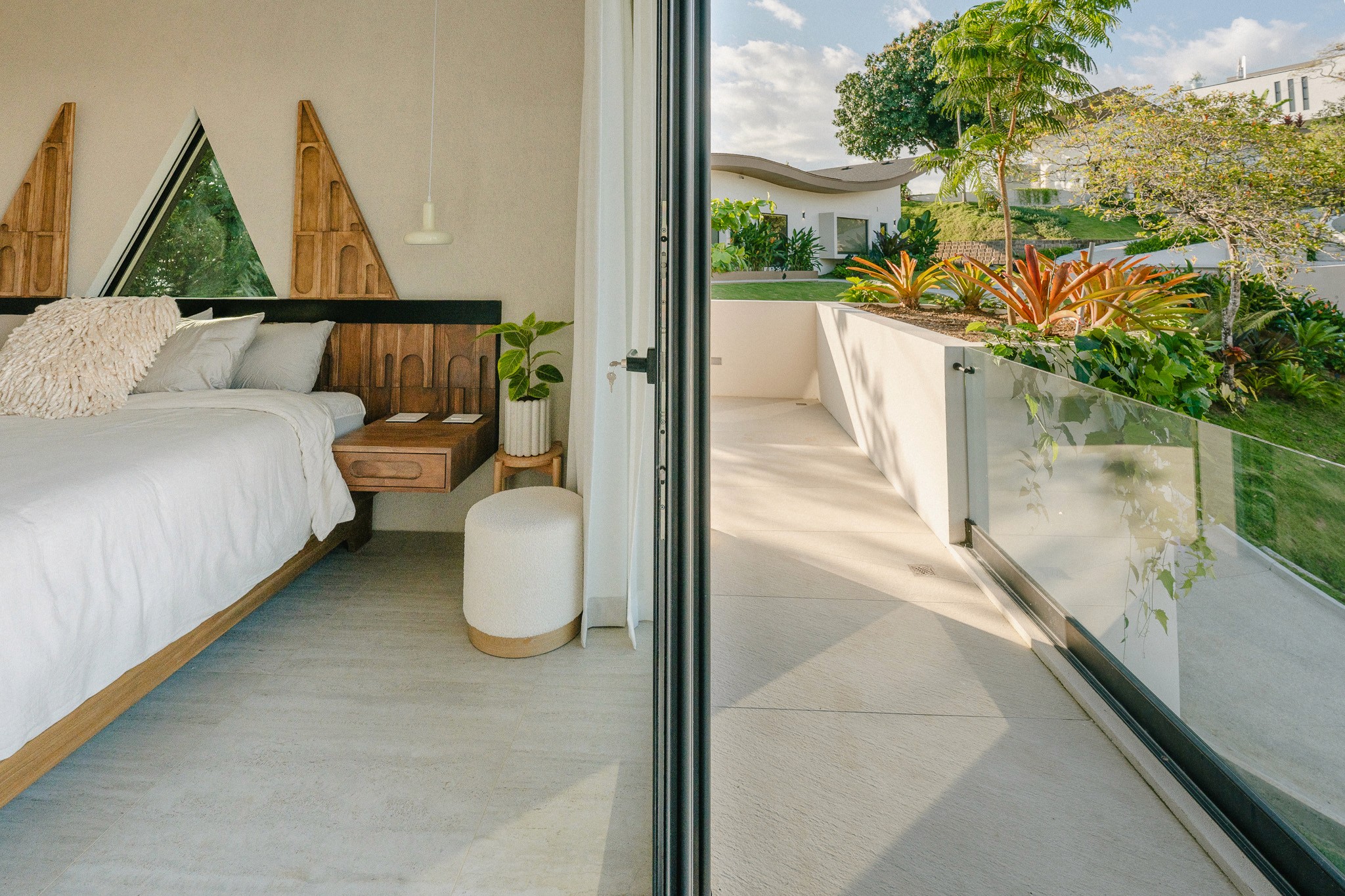Modern bedroom in a Costa Rica casita featuring a king-size bed, custom wood carved headboard, and a triangular window. Large sliding glass doors open to a private sunlit terrace with lush tropical landscaping.