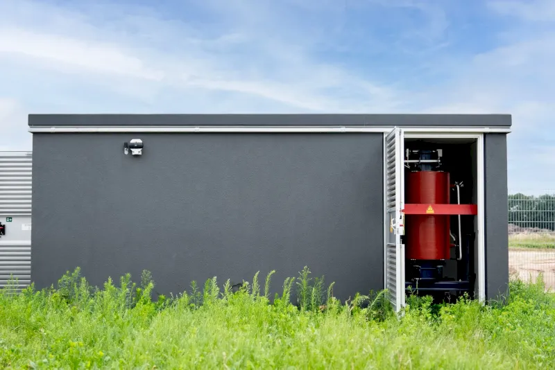 Grauer Container mit offenem Eingang, rote Maschine sichtbar, grünes Gras davor.