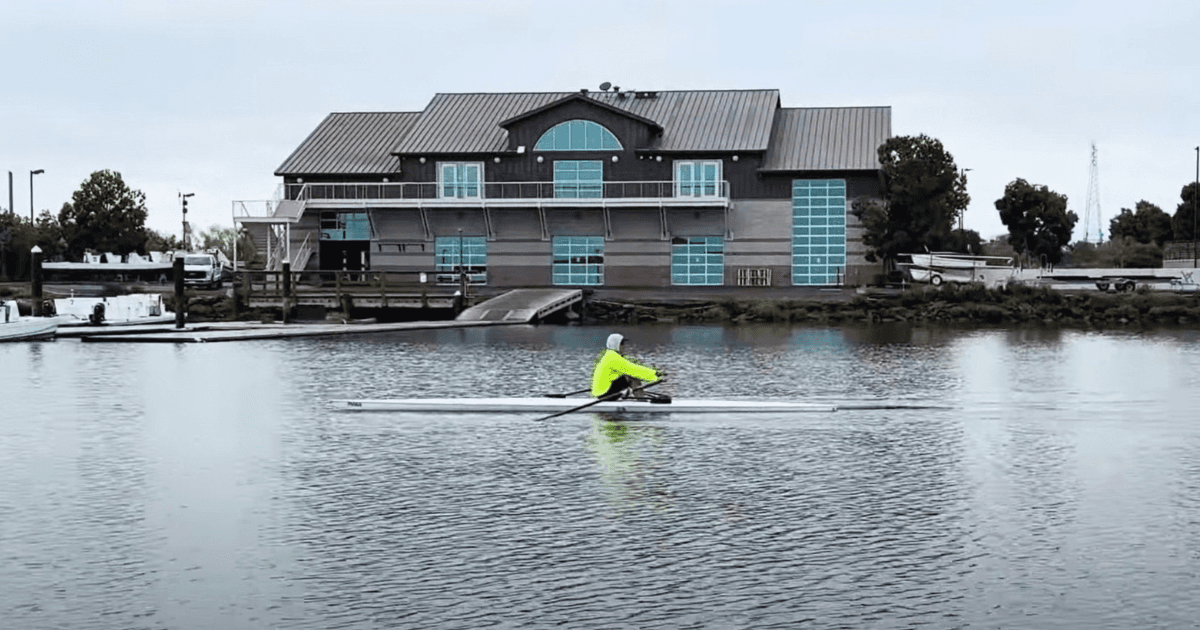Novice sculler in a yellow jacket rowing a single past a boathouse
